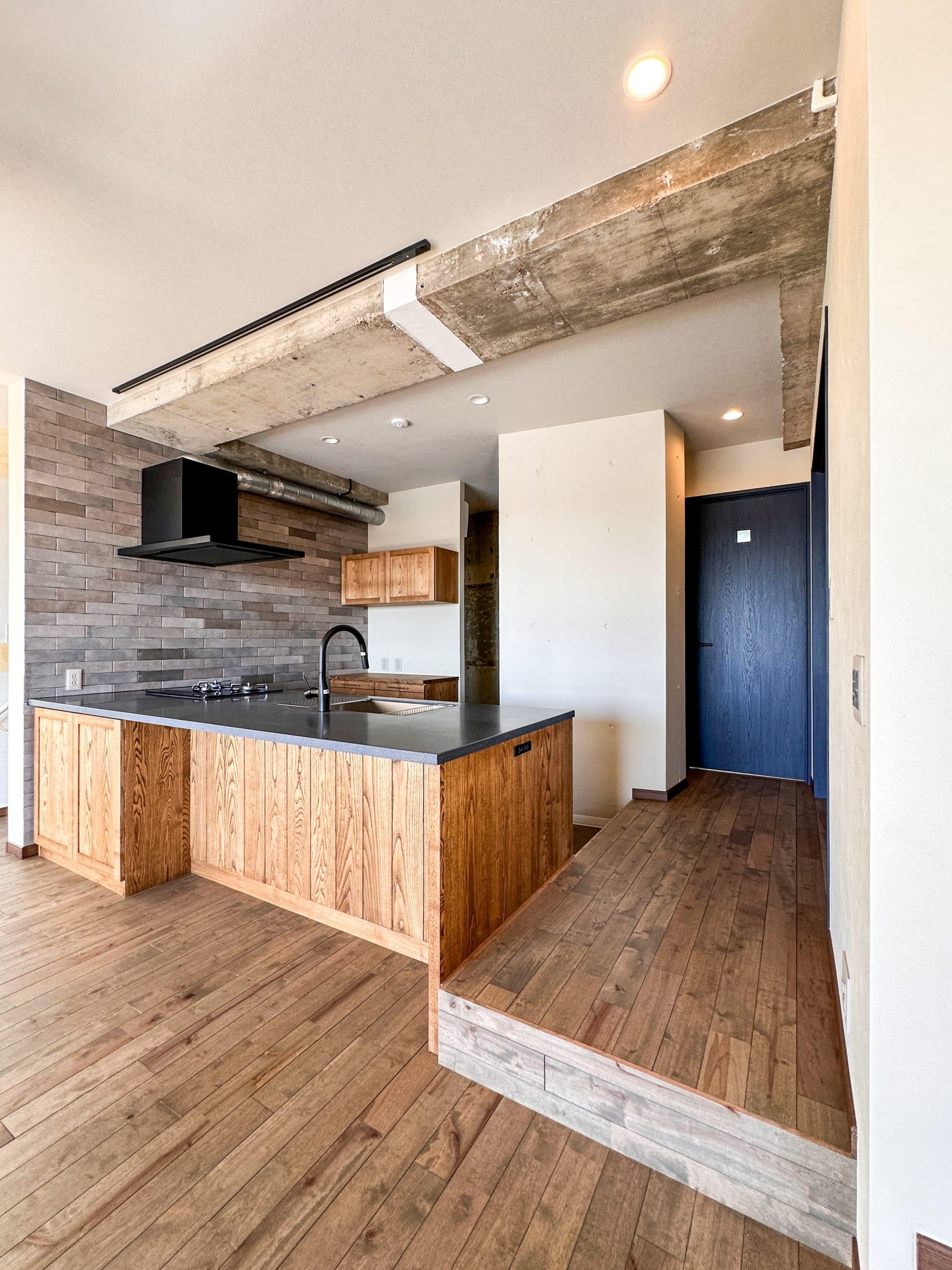 木目のキッチンとグレートーンのワークトップ、あらわしのコンクリート梁が特徴のLDK。ブラウン系の床材とタイル壁が落ち着いた雰囲気をつくる。