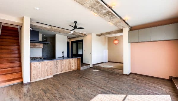 キッチン、小上がり、階段がひとつにつながるLDK全景。木の素材感とコンクリート梁のラフさが調和し、大きな窓から光が差し込む開放的な空間。