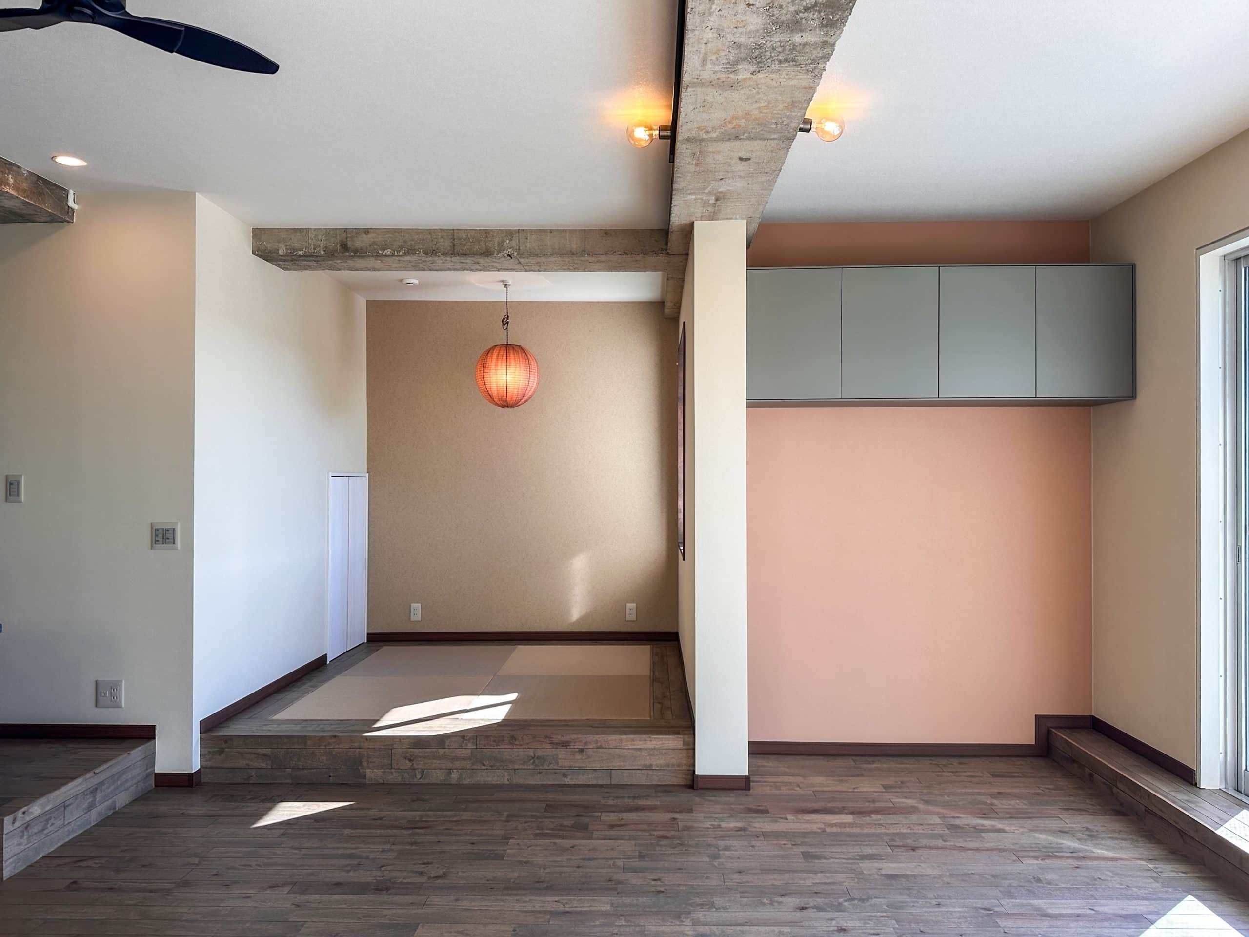 小上がりのあるリビング空間。ピンクベージュのアクセントウォールと宙に浮く造作収納、木質フロア、コンクリート梁の質感が調和したやわらかな印象の内装。